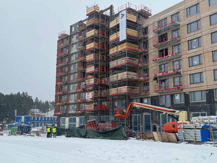 En byggarbetsplats i snöväder med en byggnad omgiven av ställningar och arbetare i närheten.