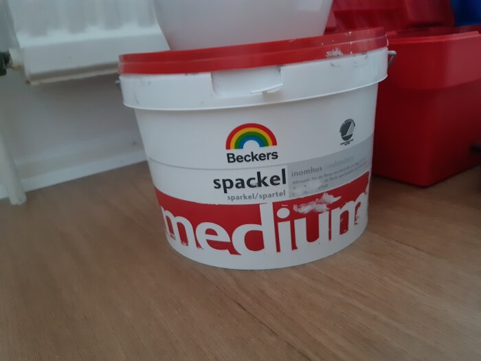 Hink med Beckers spackel, medium, använd till inomhusarbete, på trägolv nära vit och röd behållare.