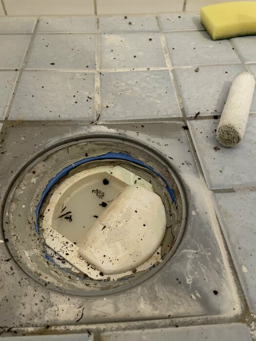 Smutsig golvbrunn med kakel, en svamp och en roller pensel i badrum.