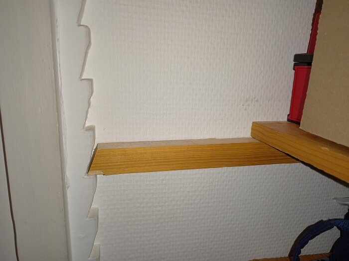 Vägg med skadad tapet, trähylla, vit dörrkarm, delvis synlig röd verktygklämma.