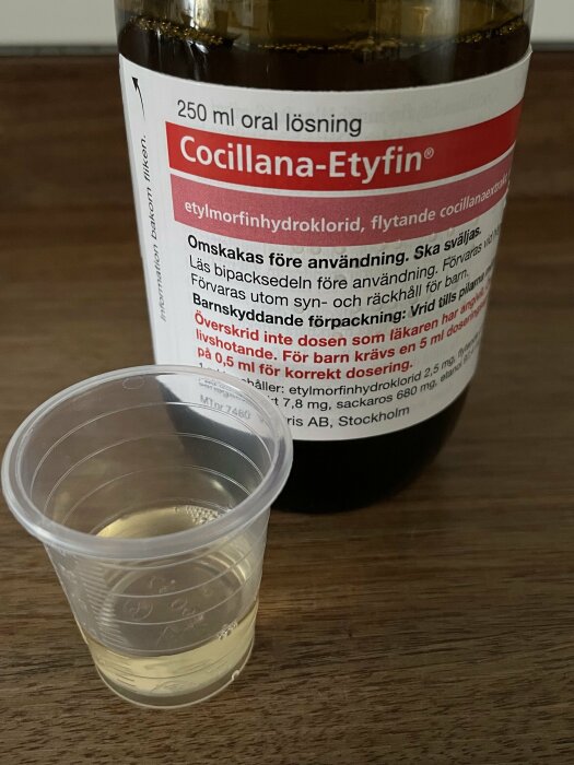 Flaska med oral lösning Cocillana-Etyfin bredvid doseringsbägare på träyta. Medicin, etikettinformation synlig.