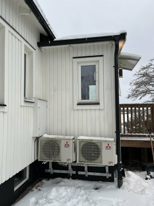 Vit trähusvägg, fönster, två luftvärmepumpar, snö, veranda i bakgrunden.