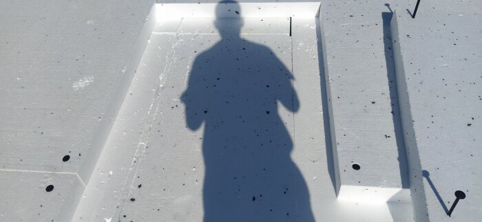 En skugga av en person speglar sig på en vit yta med små svarta prickar och linjer.