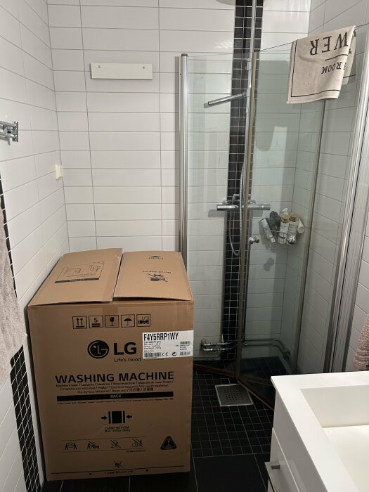 Ett badrum med vita kakelväggar, en duschhörna och en oöppnad tvättmaskinskartong från LG.