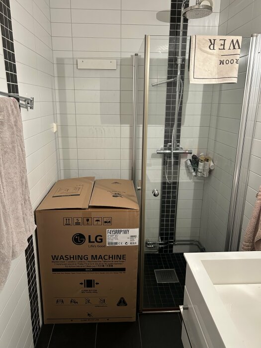 Ett badrum med en ny LG tvättmaskin i kartong, duschhörna, handduk och handfat synligt.