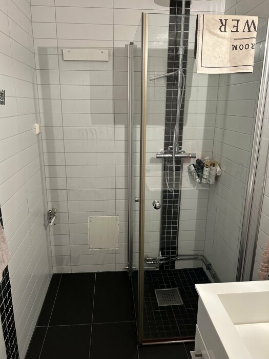 Modernt badrum, duschhörna med glasdörrar, vita kakelväggar, svart golv, handdukskrok och badrumsprodukter syns.