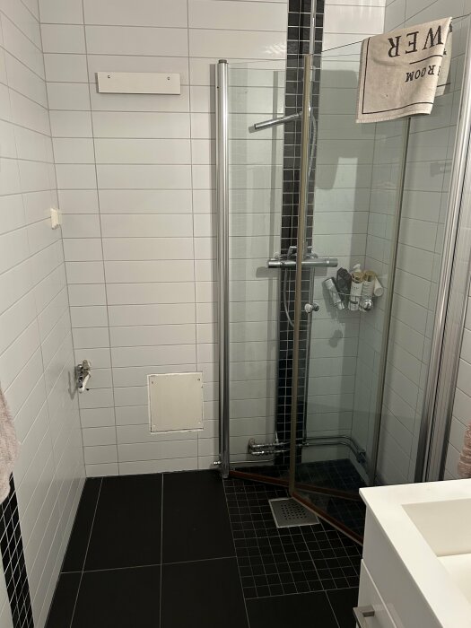 Ett modernt badrum med duschkabin, vita kakelväggar, svart golv och badrumshandduk.