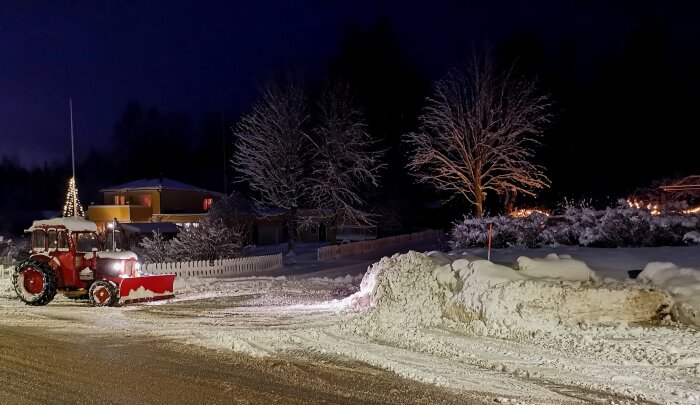 Röd traktor plogar snö nattetid, upplyst hus och träd, snöhögar, julbelysning, vinterstämning.