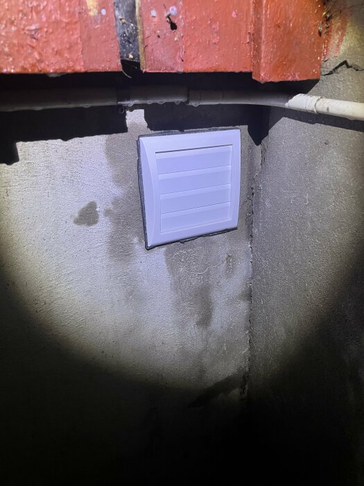 Vit ventilationsgrill på grå betongvägg under röd tegelvägg och rör. Ojämn belysning och skuggor.