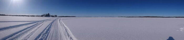 Vidsträckt snötäckt landskap med skidspår och klarblå himmel. Solen skiner starkt, och det är dagtid.