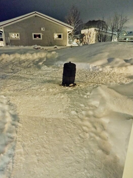 En svart soptunna på en snöig väg framför ett hus om kvällen.