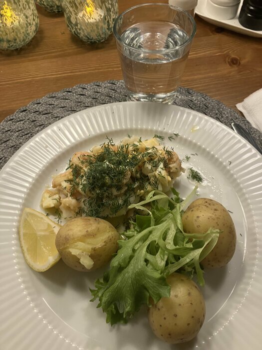 Vit tallrik med citron, potatis, sallad, fisk toppad med dill, vattenglas, träbord.