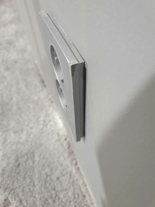 Eluttag sticker ut från vägg, dålig installation, risk för skador, säkerhetsproblem. Vit vägg, grått golv.