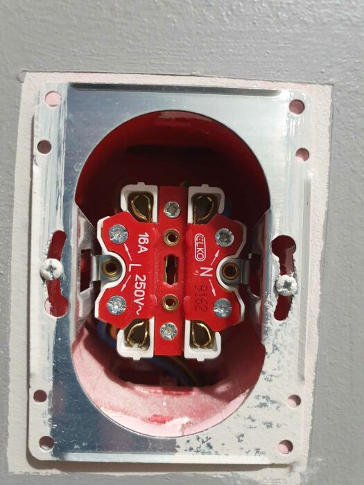 Röd eluttagsbakdel utan täckplatta monterad i vägg, märkt 16 A, 250V. Oavslutad elektrisk installation.