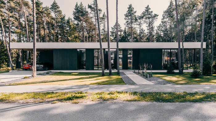 Modernt hus med platt tak, inbäddat i skogsmiljö, stora fönster, minimalistisk design, carport med bil.