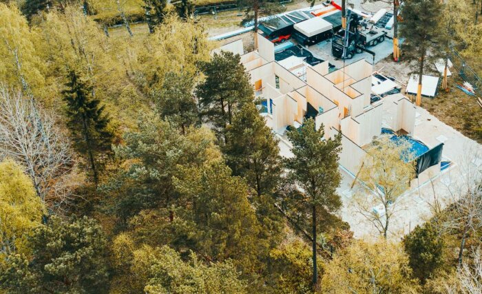 Flygbild över byggarbetsplats i skogsmiljö med pågående huskonstruktion och byggfordon.