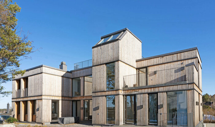 Modernt hus med träbeklädnad, stora fönster, balkonger, blå himmel, omgivet av natur.