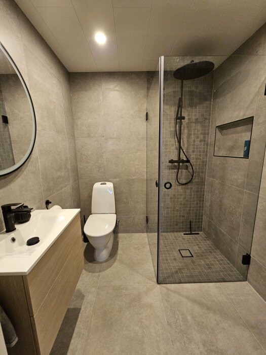 Modernt badrum, duschhörna, toalett, handfat, stor spegel, grå klinker, brunt badrumsskåp, inbyggda hyllor.
