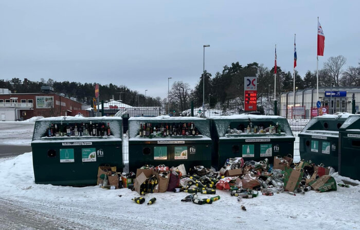 Överfulla återvinningsstationer för glas, flaskor på marken, vinter, snö, flaggor i bakgrunden, dimmig dag.