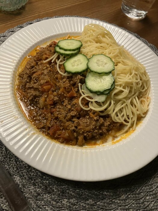 Spaghetti med köttfärssås och skivade gurkor på en vit tallrik.