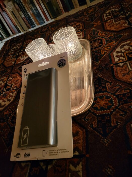 En powerbank i förpackning, två ljusglas och en silverbricka på ett orientaliskt mönstrat mattgolv framför bokhylla.