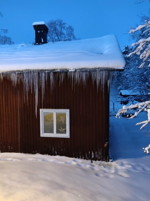 Snötäckt hus med is istappar och djupt snölager under skymning.