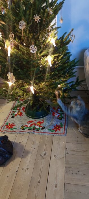 Julgran med ljus och dekorationer, katt leker, trägolv och julmatta, mysig hemmiljö.