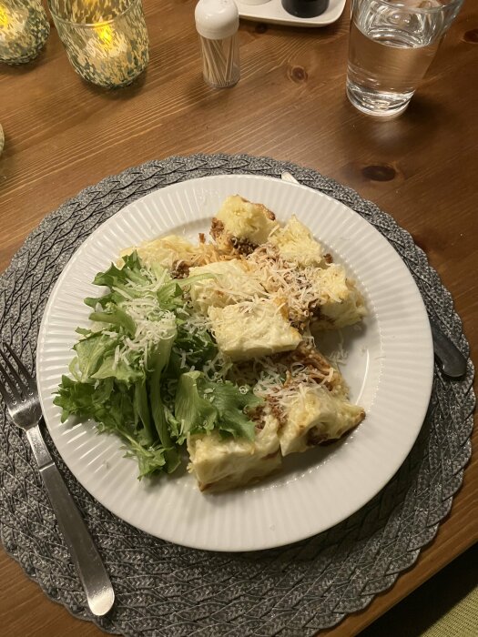 En tallrik med lasagne och grönsallad, riven ost ovanpå, bord dukat för måltid, vattenglas och saltkar.