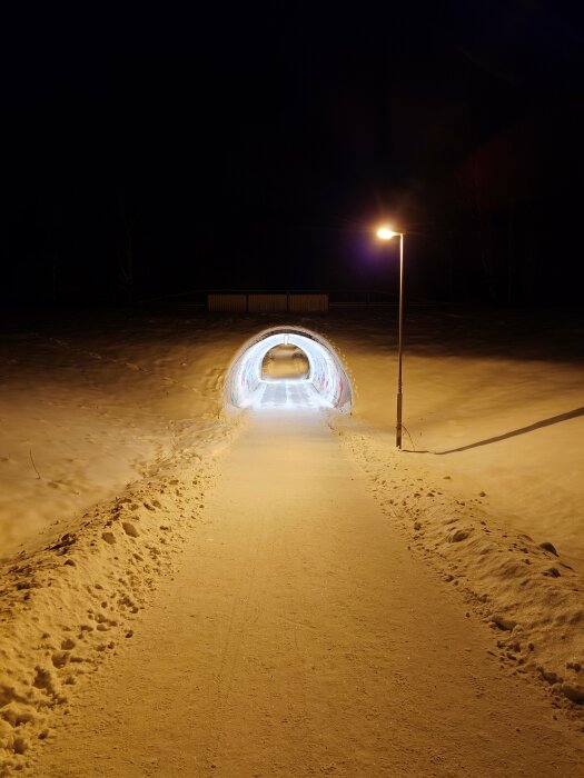 Natt, upplyst gångväg leder till en snötunnel med graffiti, gatlykta, omgivet av mörker och snö.