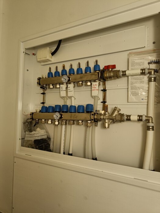 Värmefördelningsskåp för vattenburen golvvärme med flera manometrar och justeringsventiler. Dokumentation på väggen.