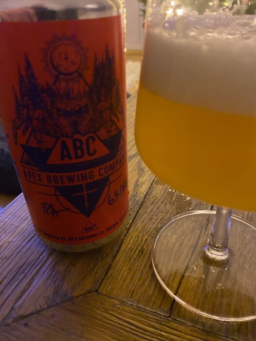 En burk IPA-öl från Apex Brewing Company intill ett glas med skum på en träyta.