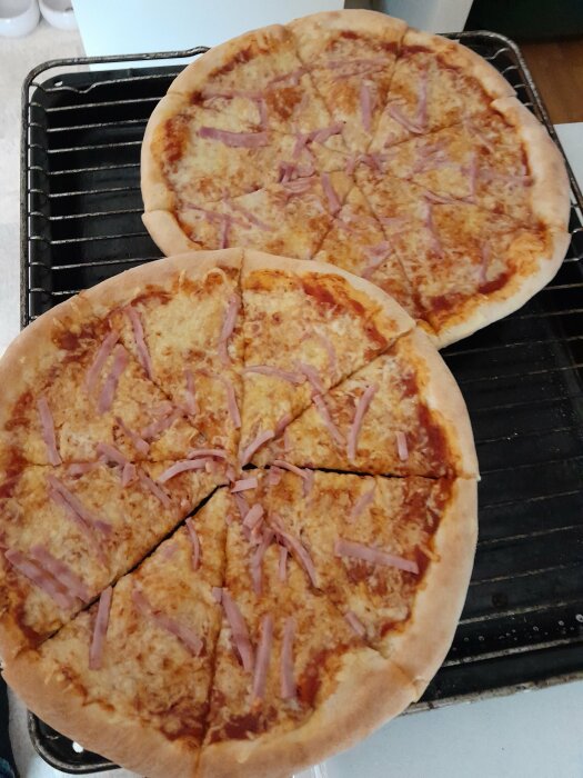 Två nystekta pizzor med skinka, uppskurna i bitar, på en ugnspanna och ett galler.