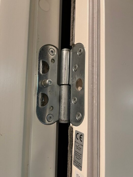 Dörrgångjärn i närbild på vit dörrkarm, synlig CE-märkning, grå stål, skruvar, genomsläppsljus.