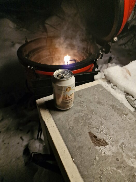 En ölburk på ett snötäckt bord vid en grill i mörker. Lågor syns inuti grillen.