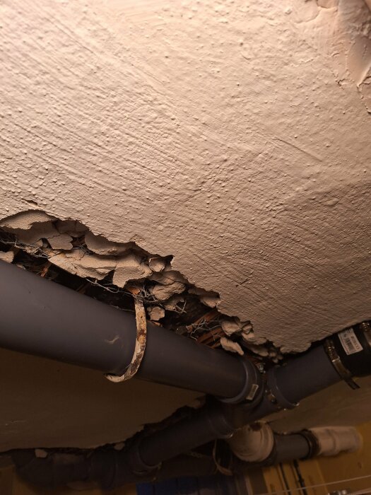 Skadad takgips runt rörledningar, exponerade lager och installationer, behov av reparation.