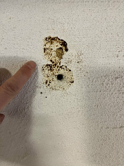 Finger pekar på brun fläck på vit texturerad vägg. Möjlig korrosion eller smuts.