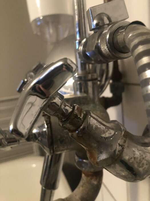 Närbild av en korroderad duschmunstycke och slang mot en kaklad vägg i badrum.