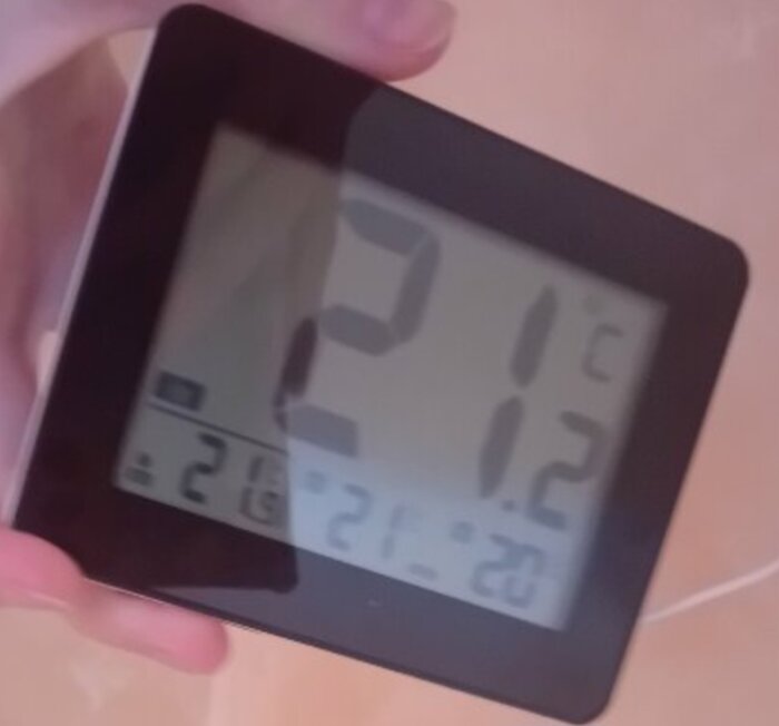 Hand håller digital termometer som visar temperaturer i Celsius; inomhus- och utomhusvärden med datumfunktion.