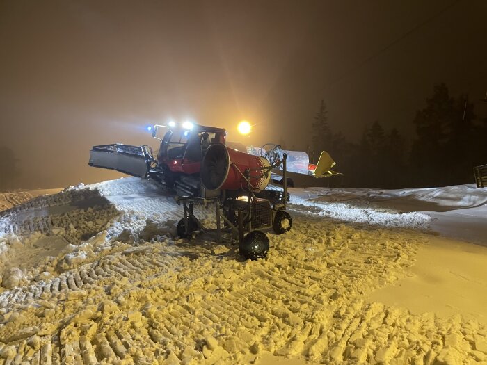 Pistmaskin i arbete nattetid, preparerar snö för skidåkning, starka strålkastare, dimmig bakgrund.