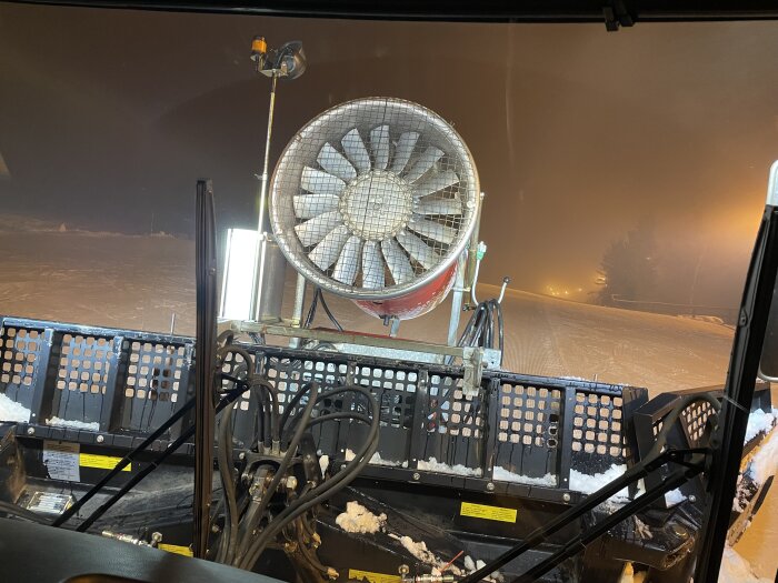 Inifrån en pistmaskin som preparerar en skidbacke nattetid med snökanoner och belysta sluttningar i bakgrunden.