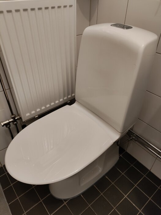 Vit toalettstol, toalettlock öppet, svartvit klinkergolv, vit kakelvägg, vit radiator till vänster.