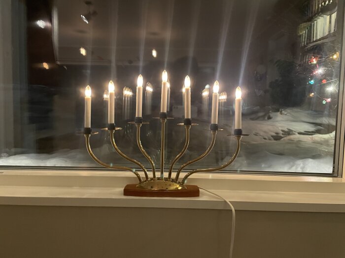Menorah med tända ljus i fönster, reflektioner, snötäckt landskap och julgransljus ses utanför.
