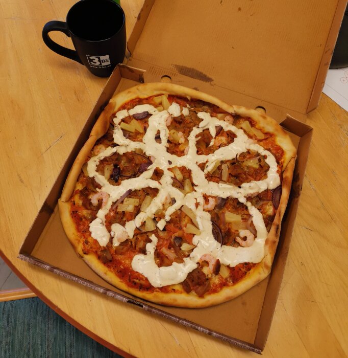 En pizza med olika pålägg och vit sås i kartong bredvid en svart mugg.