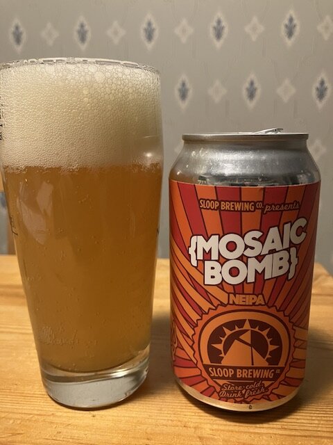 Ölglas med skummande öl bredvid en burk märkt "Mosaic Bomb! NEIPA" från Sloop Brewing Co.