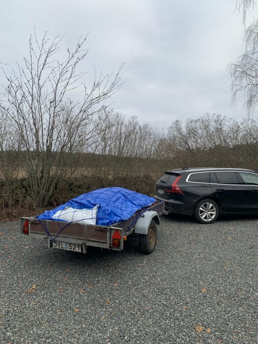 Svart bil kopplad till släpvagn täckt med blå presenning, på grusparkering, trista vinterträd i bakgrunden.