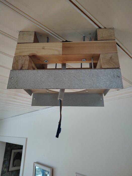 Osäker takmontering av trä och metall med avskalad elektrisk ledning hängande ner.
