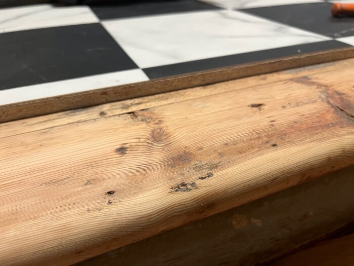 Träkant i förgrunden, suddig svartvit schackrutig bakgrund, inomhus, naturliga material, byggmiljö, detaljnärbild.
