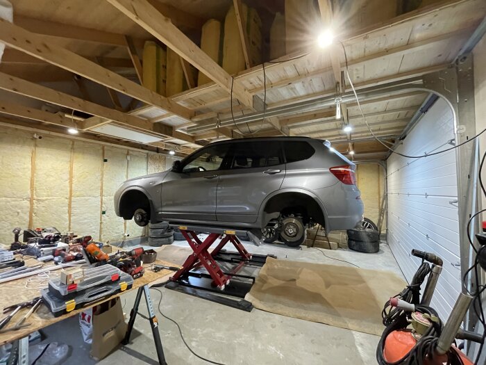 Garage med SUV på lyft, verktyg på bord, oorganiserat, däck och verkstadsutrustning synliga.