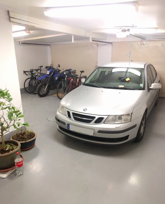 Vit bil parkerad i garage med cyklar, plantor och en Coca-Cola-flaska på golvet.