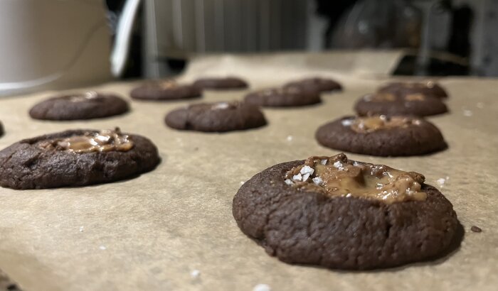 Nybakade chokladkakor med smält fyllning och flingsalt på bakplåtspapper, köksmiljö i bakgrunden.